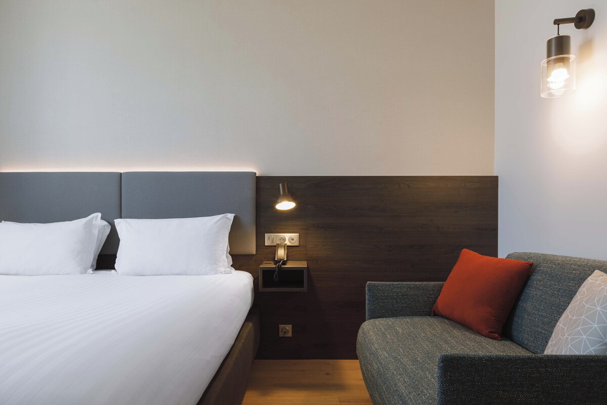 Habitación de hotel con cama, mesita de noche con lámpara y sofá con cojines.
