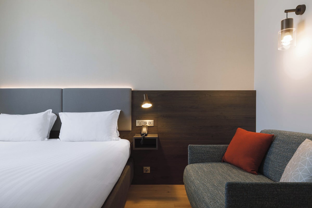 Habitación de hotel con cama, mesita de noche con lámpara y sofá con cojines.