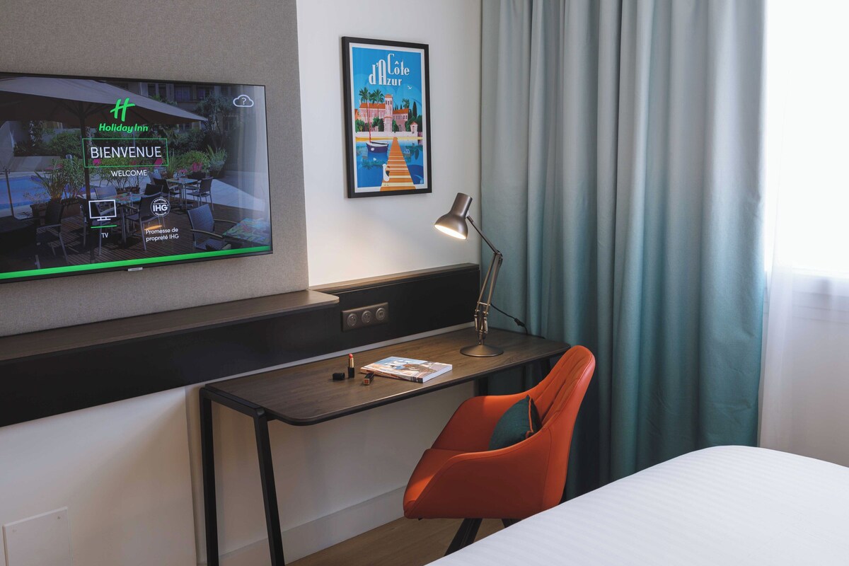 Habitación de hotel con una televisión mostrando una pantalla de bienvenida, un escritorio con lámpara, una silla y una cama.