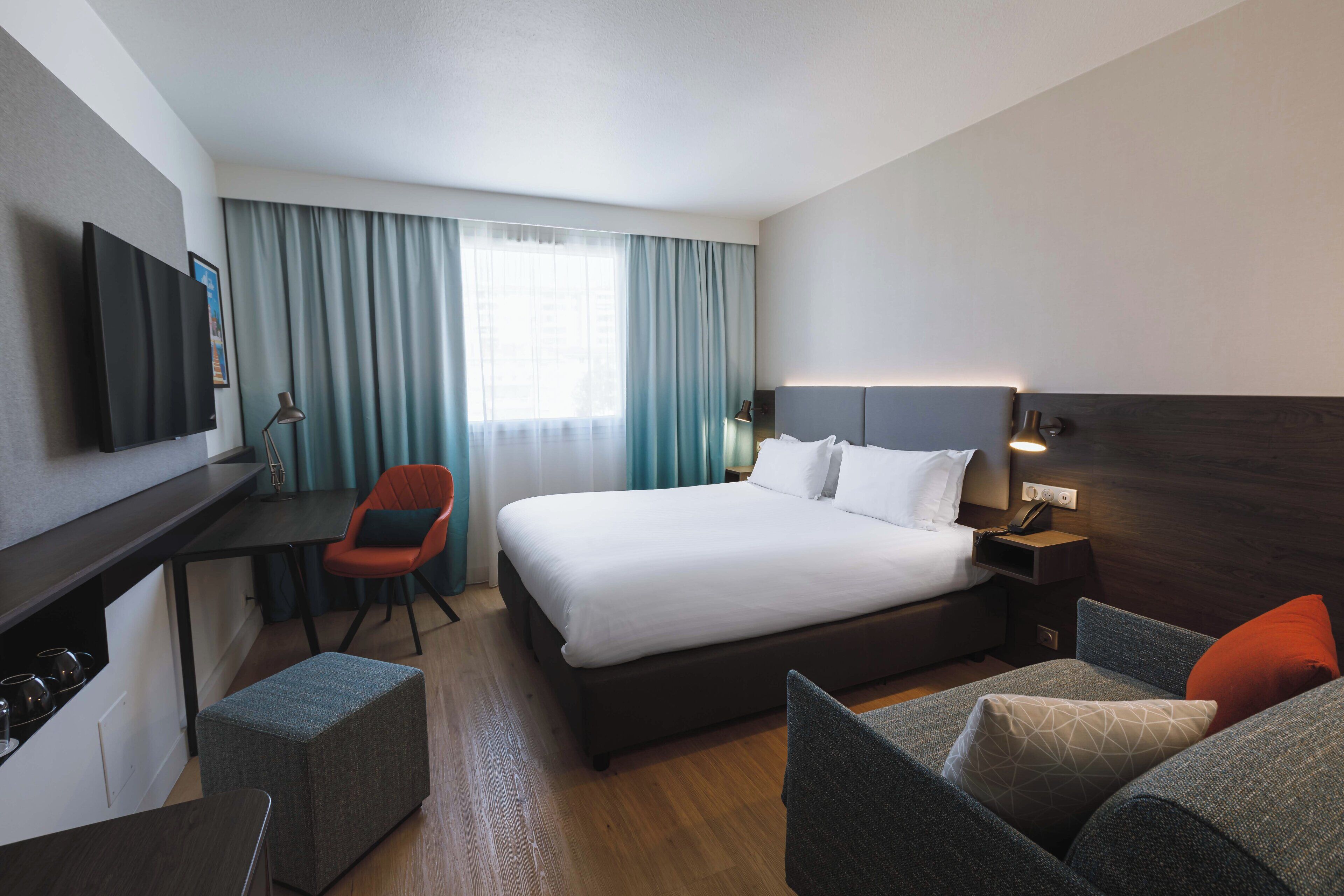 Ein Hotelzimmer mit einem großen Bett, einem Schreibtisch mit Stuhl, einem an der Wand befestigten Fernseher und einem Fenster mit Vorhängen.