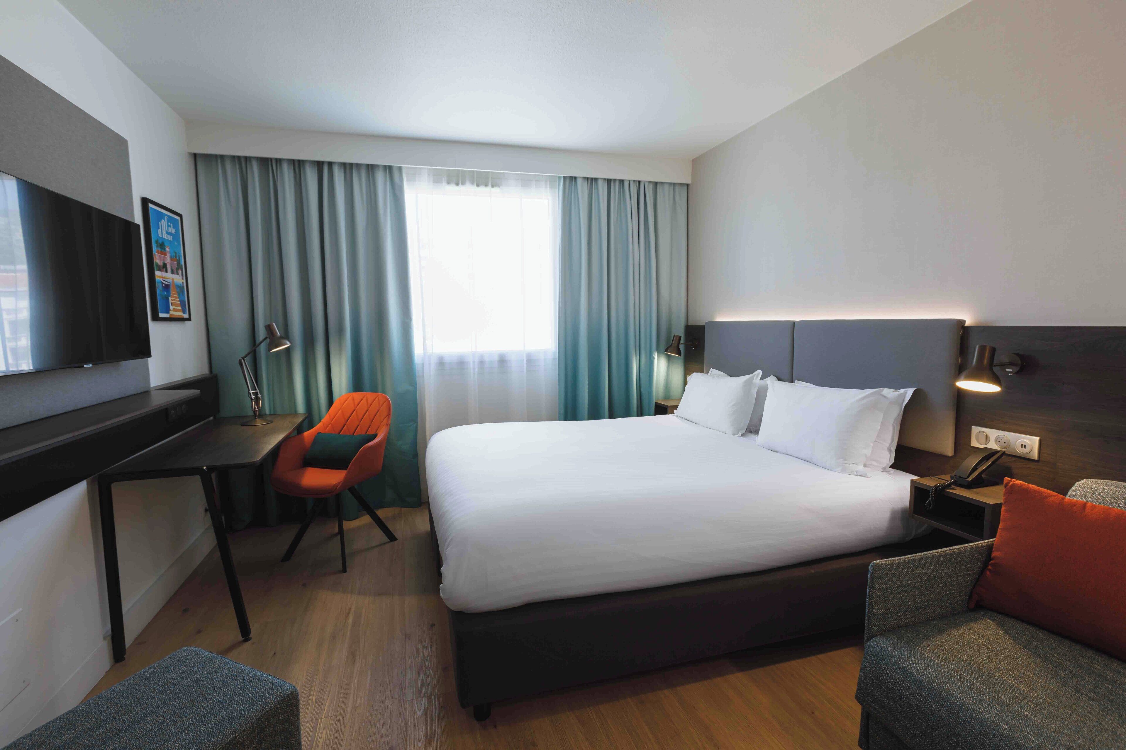 A hotel room with a large bed, a desk with a chair, a TV, and a window with curtains.