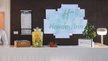 A reception desk with a sign for Holiday Inn, a tariffs room rates poster, a jar of lemons, a flower, and a sign for IHG One Rewards.