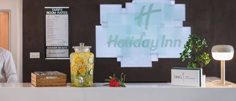A reception desk with a sign for Holiday Inn, a tariffs room rates poster, a jar of lemons, a flower, and a sign for IHG One Rewards.