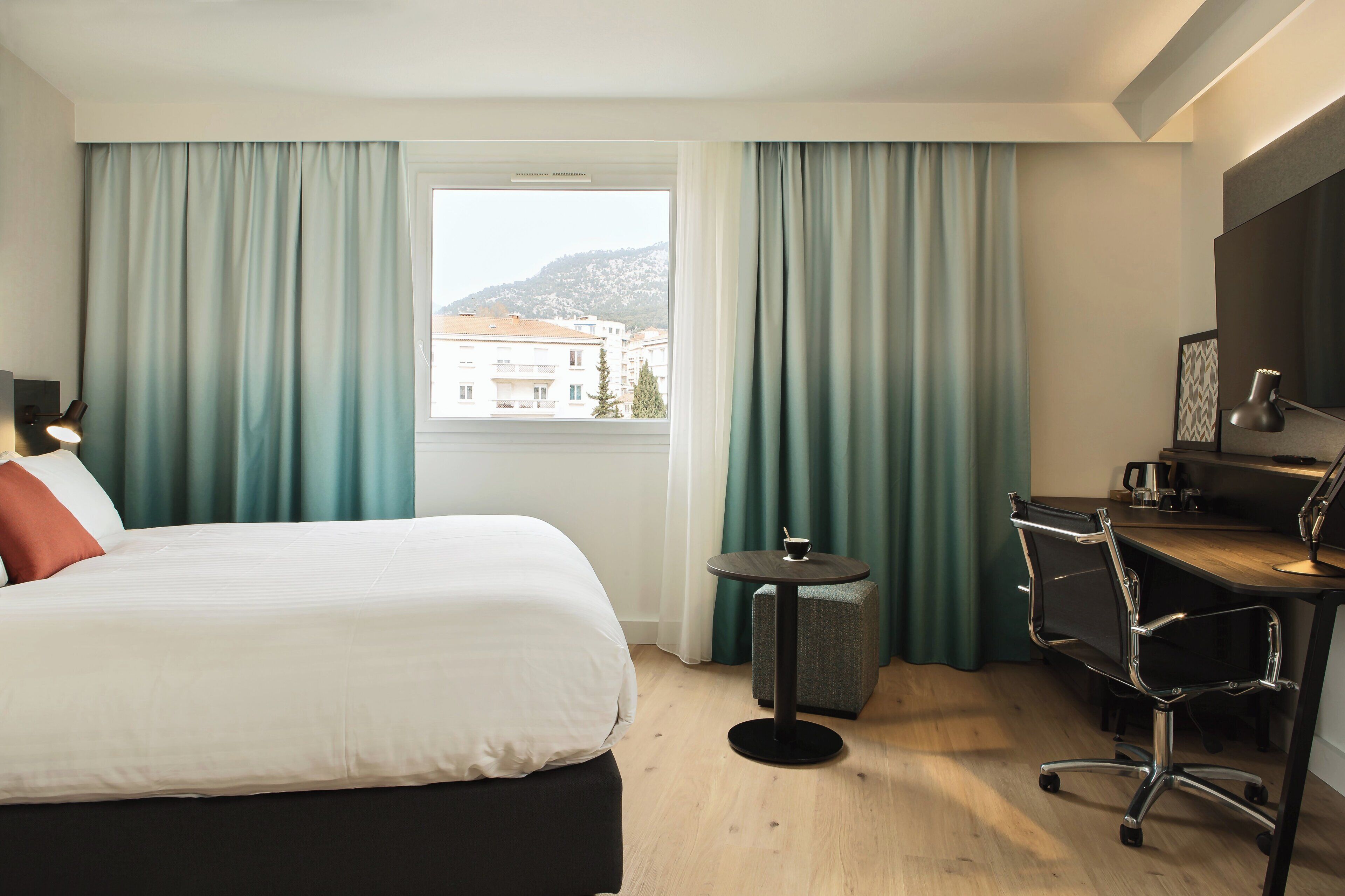 A hotel room with a bed, a desk, a chair, and a view of buildings through the window.