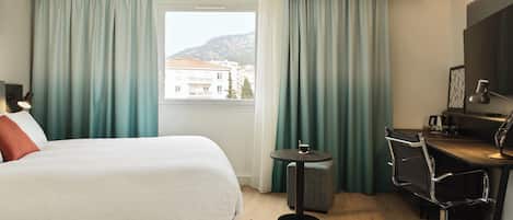 A hotel room with a bed, a desk, a chair, and a view of buildings through the window.