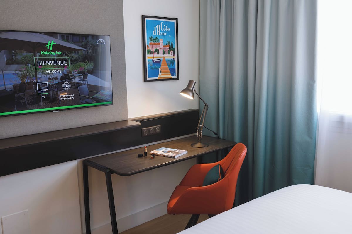 Habitación de hotel con una televisión mostrando una pantalla de bienvenida, un escritorio con lámpara, una silla y una cama.