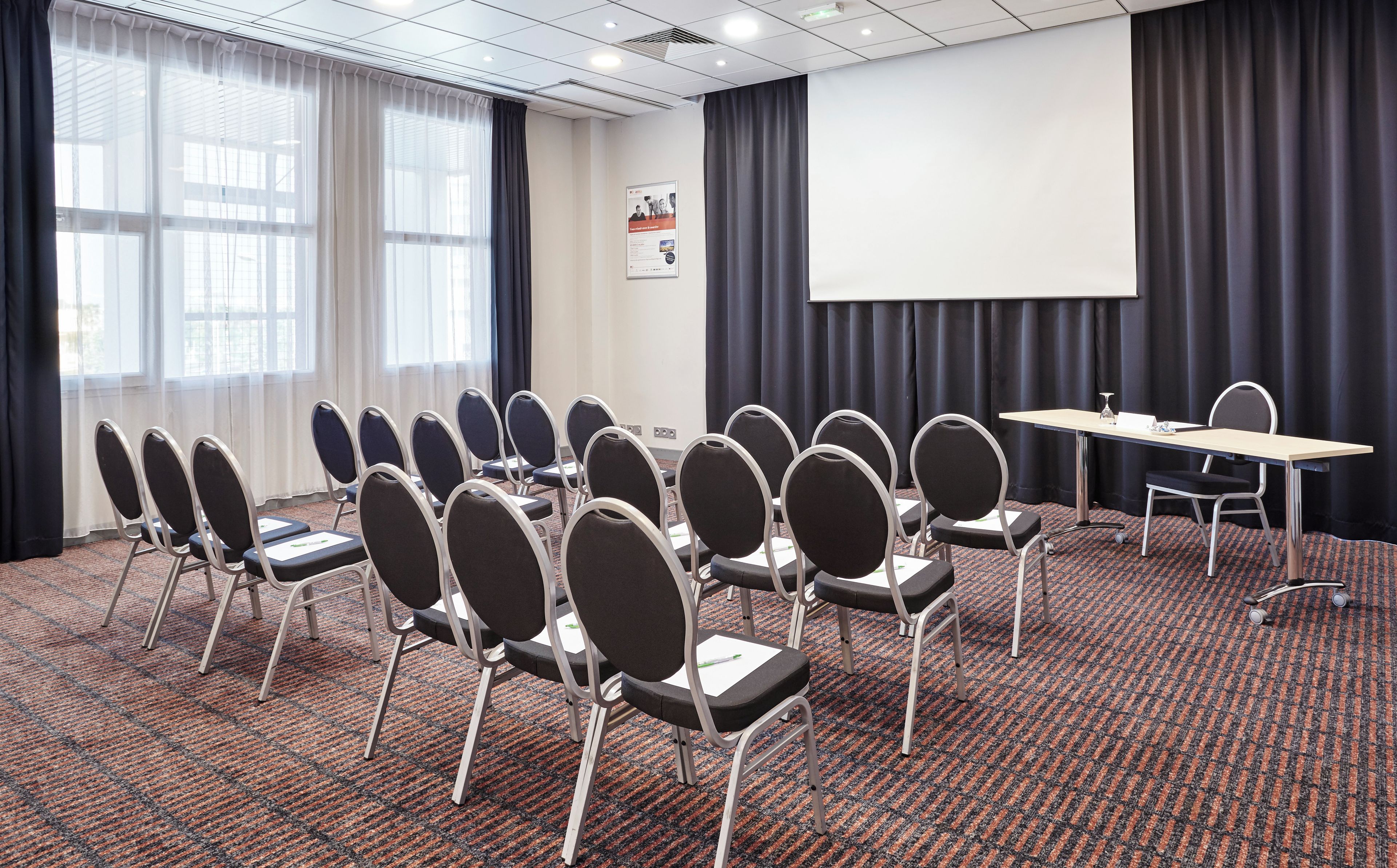 Une salle de conférence avec des rangées de chaises, un écran de projection et un sol recouvert de moquette.