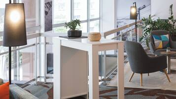 A modern hotel room with a white desk, a grey armchair, a potted plant, and a large window offering a city view.