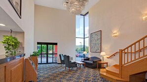 Una hall con una scala in legno, una reception, un tappeto blu decorato, un lampadario e un'ampia finestra con vista sull'esterno.