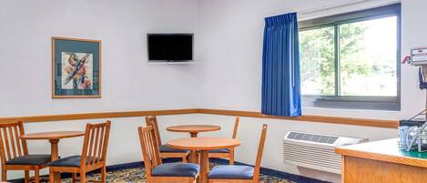 A hotel room with a corner seating area, a flat-screen TV mounted on the wall, a window with blue curtains, and a patterned carpet.