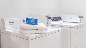 Una lavatrice e asciugatrice Whirlpool a carica frontale in una lavanderia bianca e pulita, con asciugamani piegati con cura e una scatola di detersivo All sopra la lavatrice.