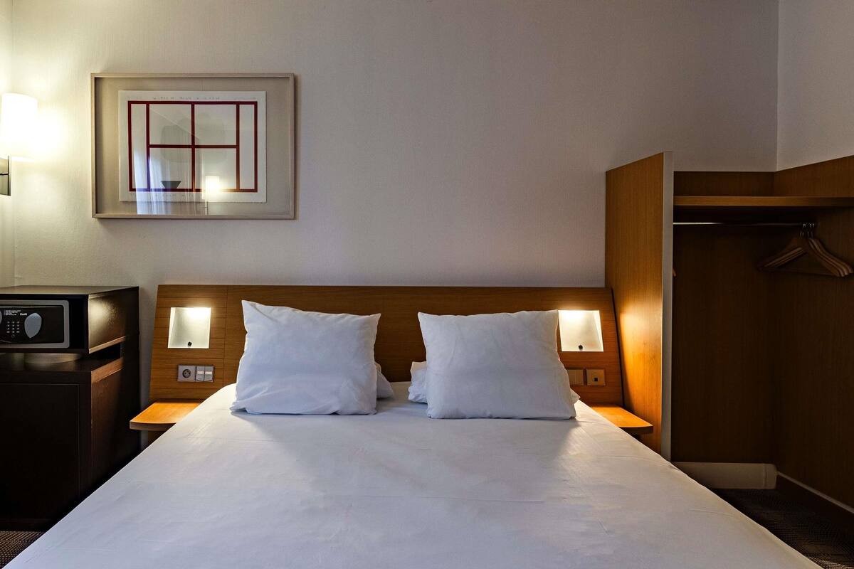 A hotel room with a large bed, two bedside lamps, a wooden headboard, a framed picture on the wall, a radio on the nightstand, and a coat rack.
