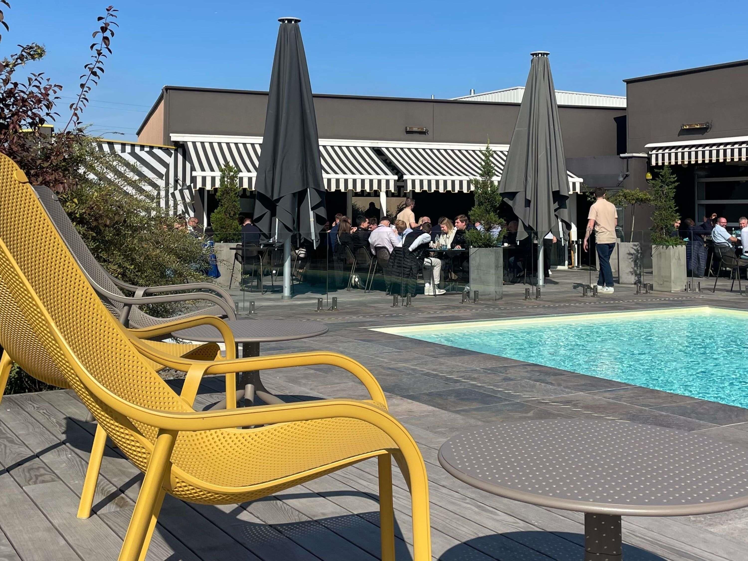 Un espace aménagé au bord de la piscine, avec des chaises longues jaunes, une piscine et, au fond, un bâtiment doté d’auvents à rayures.