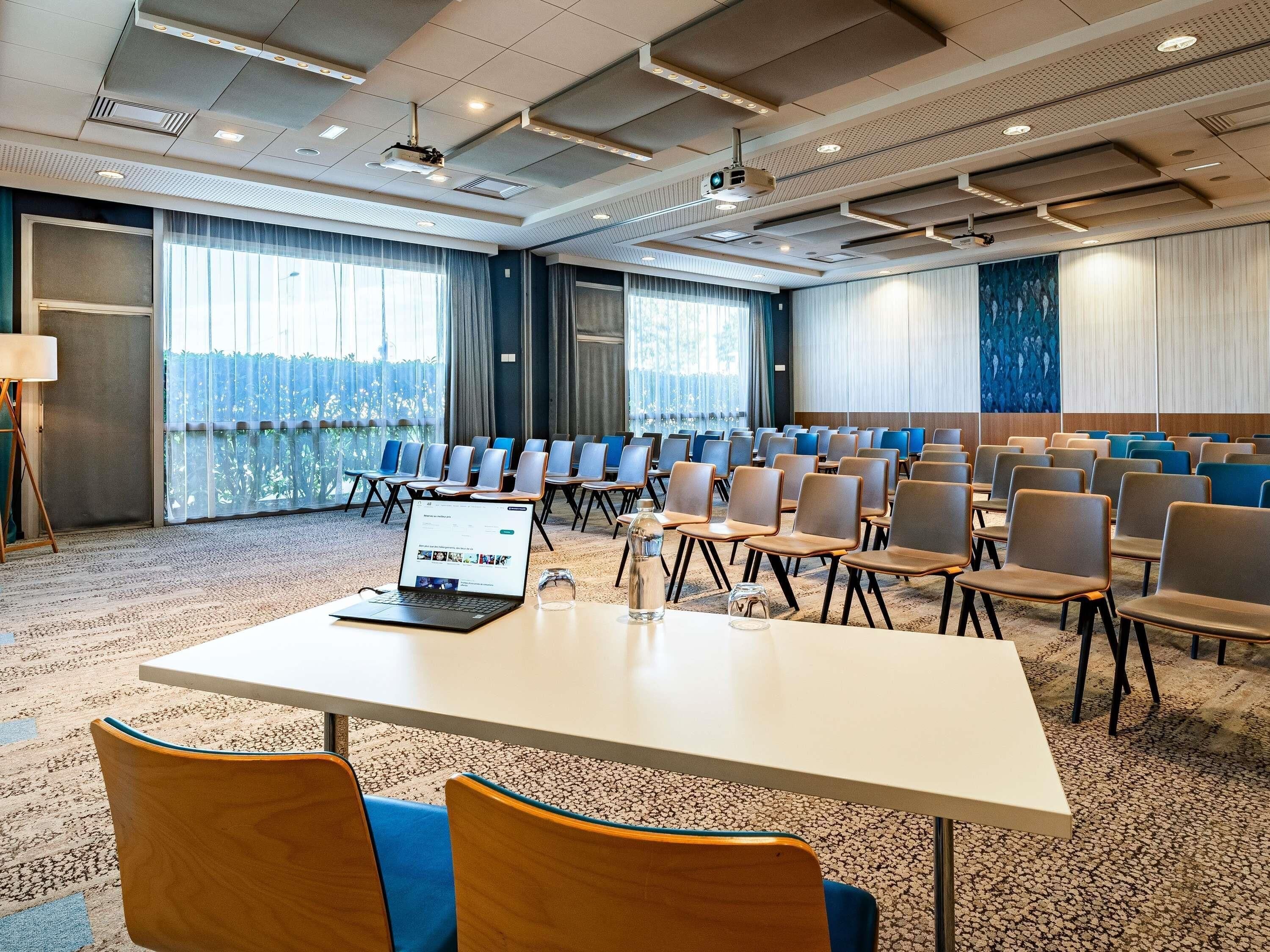 A conference room with rows of chairs, a laptop on a table, and large windows with curtains.
