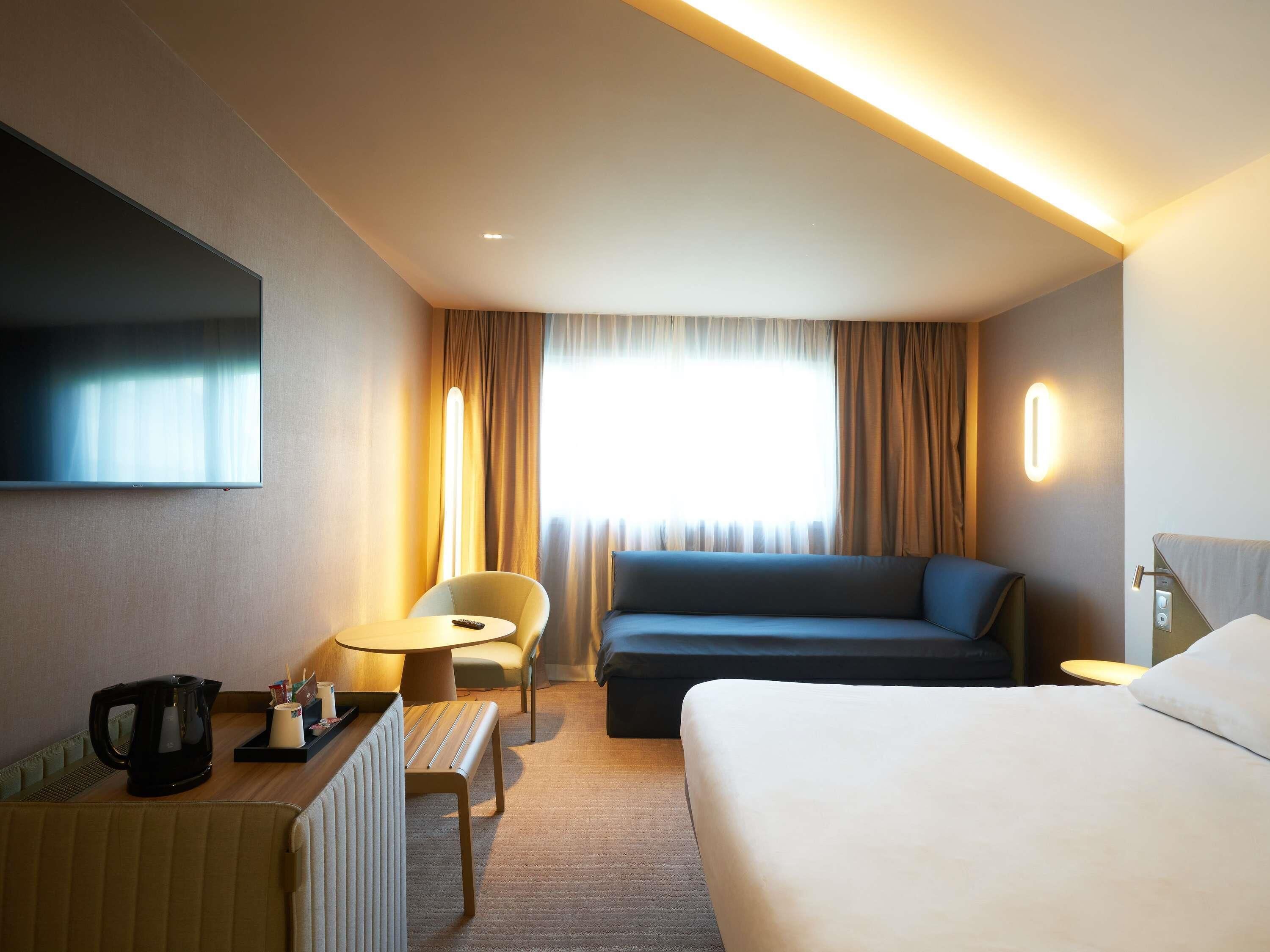 Ein Hotelzimmer mit einem Bett, einer Couch, einem Sessel, einem kleinen Tisch mit Wasserkocher und einem an der Wand befestigten Fernseher.