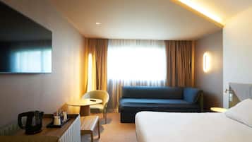 Habitación de hotel con cama, sofá, sillón, una mesita con una tetera y un televisor en la pared.