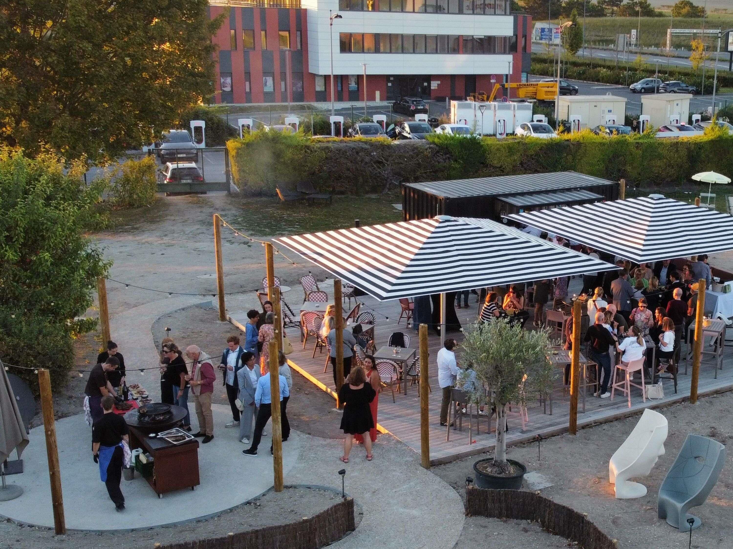 Un área de comedor al aire libre con mesas y sillas, rodeada de follaje y un edificio moderno al fondo.