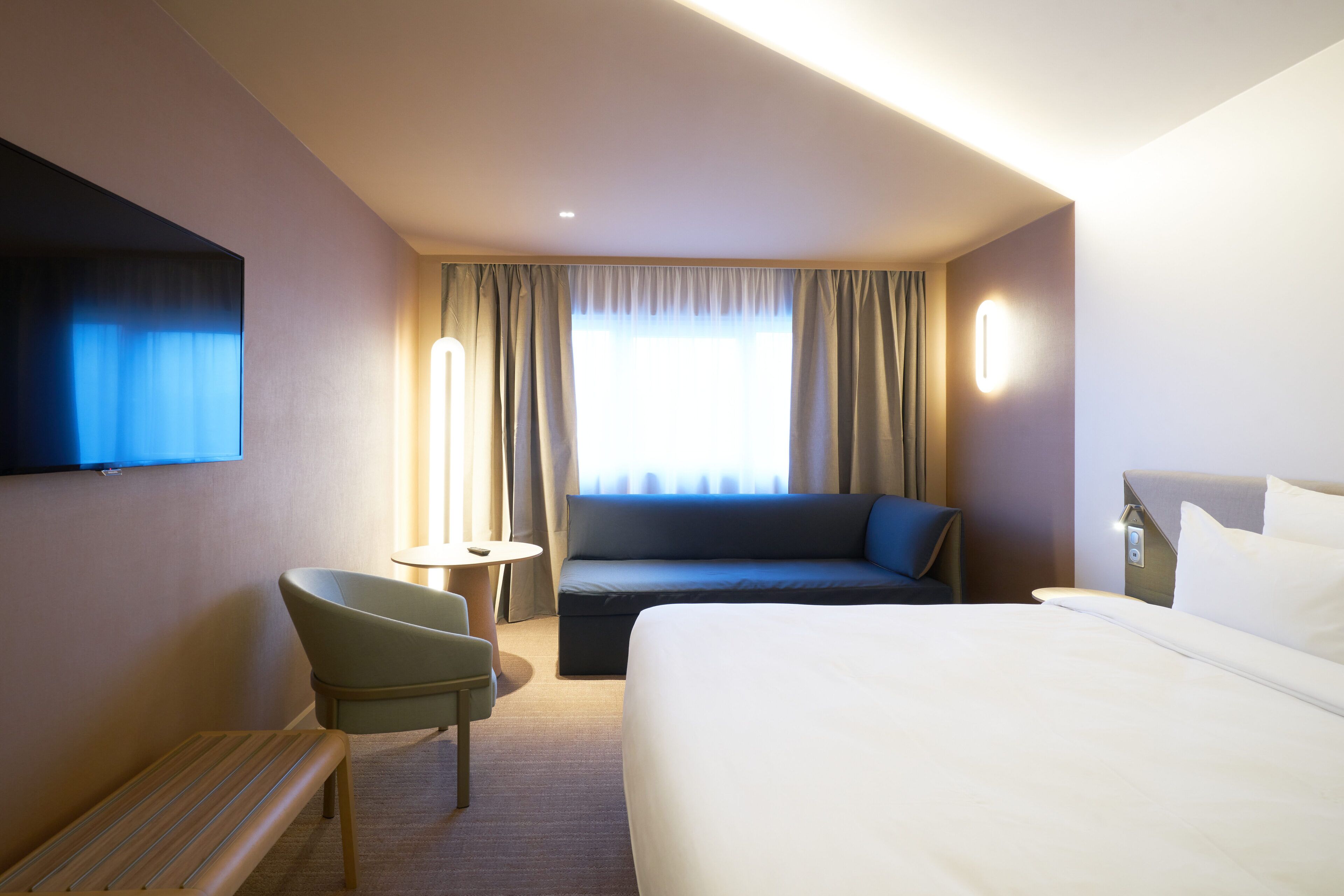 A hotel room with a bed, a sofa, a chair, a small table, and a TV mounted on the wall.