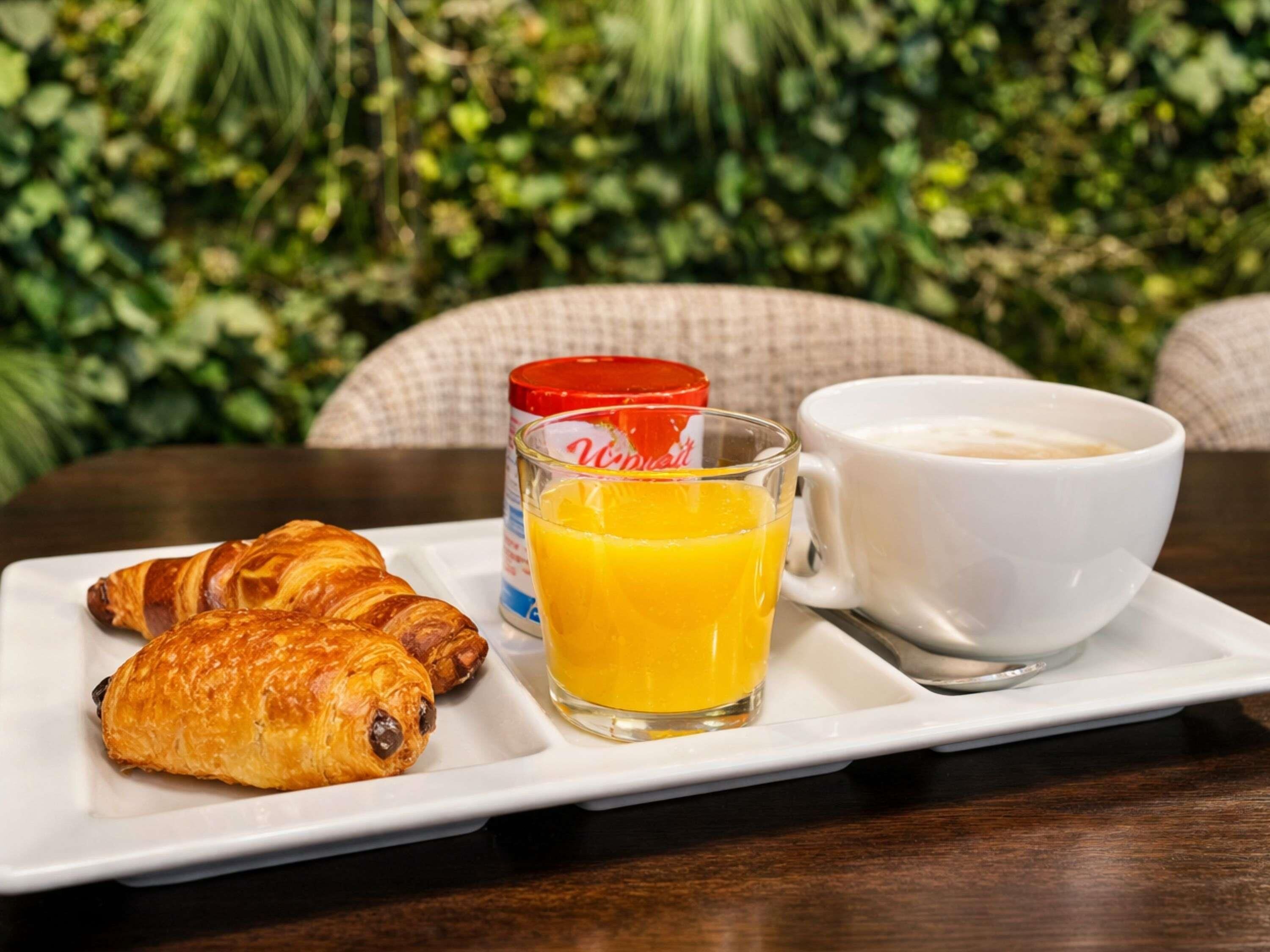 A breakfast tray with croissants, orange juice, and a cup of coffee on a table with a green leafy background.