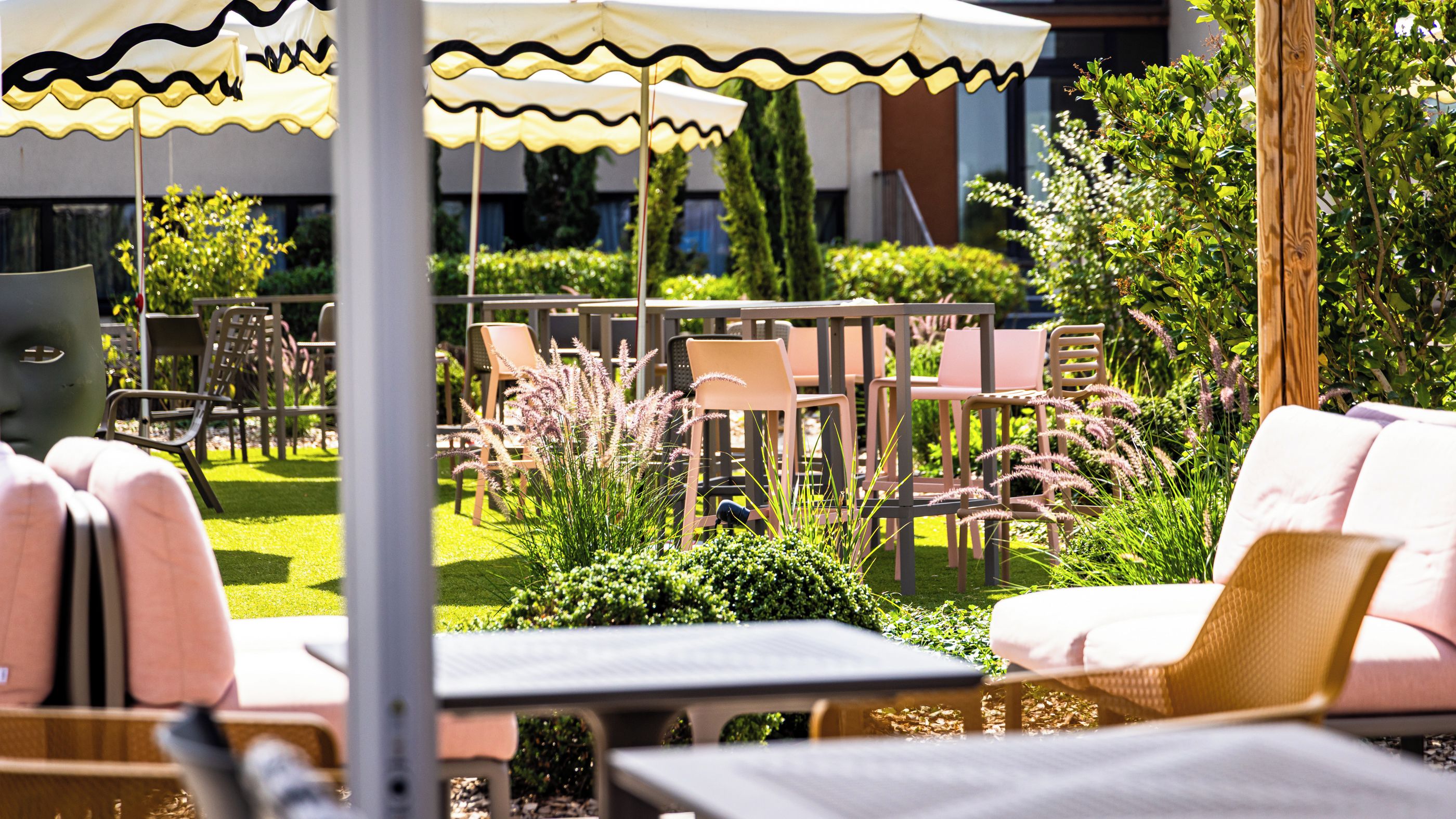 An outdoor seating area with tables and chairs, surrounded by greenery and a building in the background.