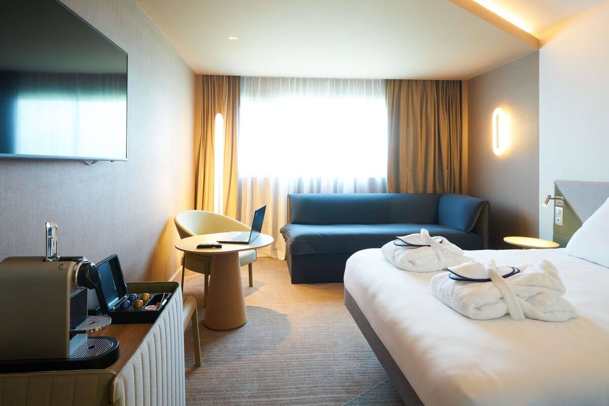 A hotel room with a bed, a sofa, a desk with a laptop, a coffee machine, and a TV mounted on the wall.