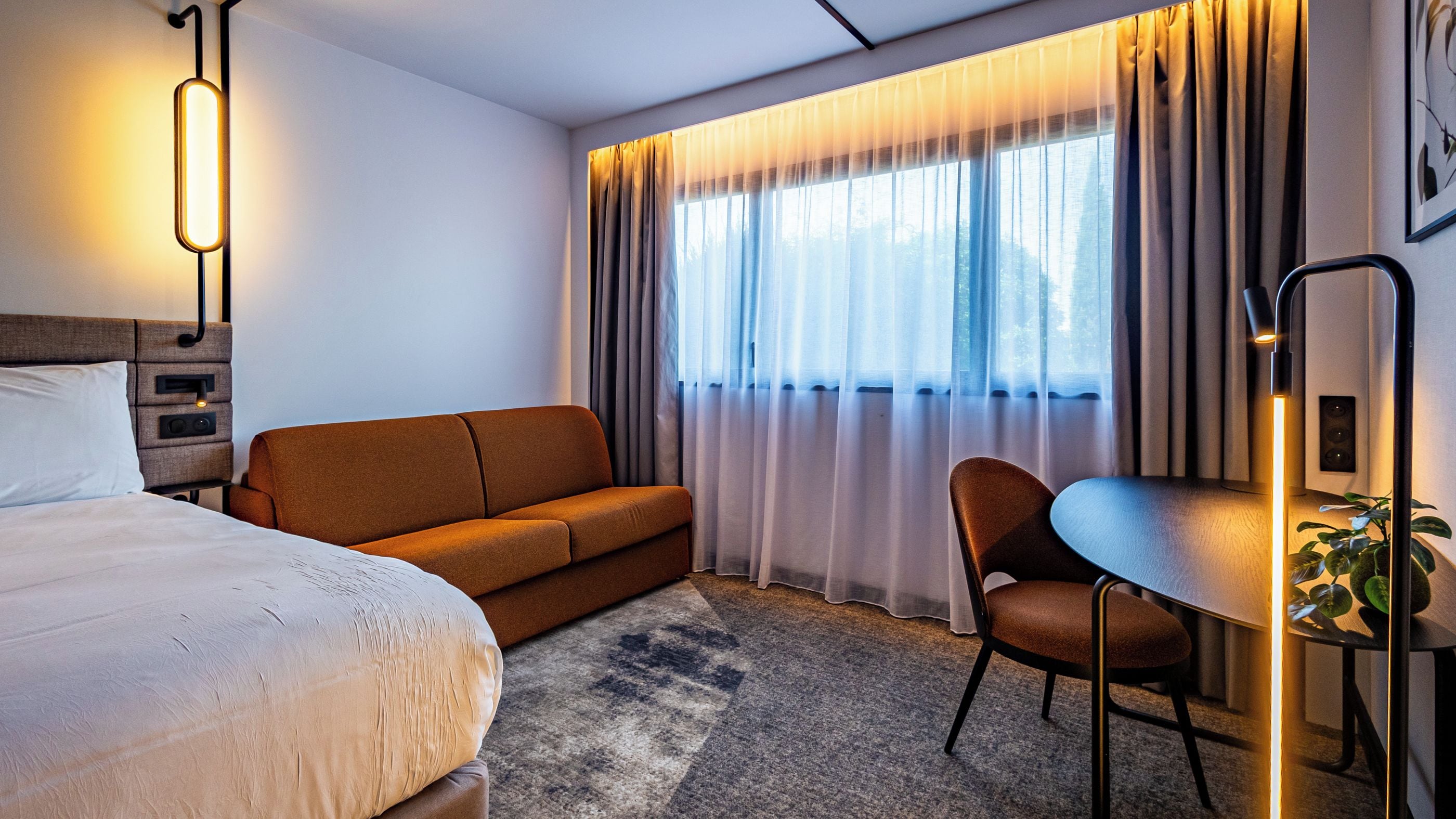 Habitación de hotel con cama, sofá, sillón, mesa y ventana con cortinas.