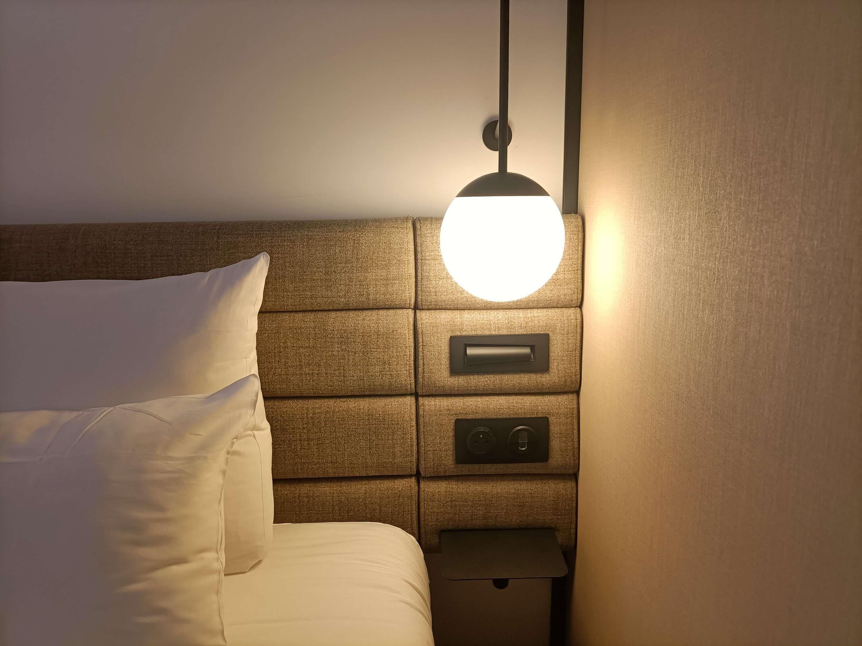 A hotel room with a bed, a bedside table with a lamp, and a control panel.