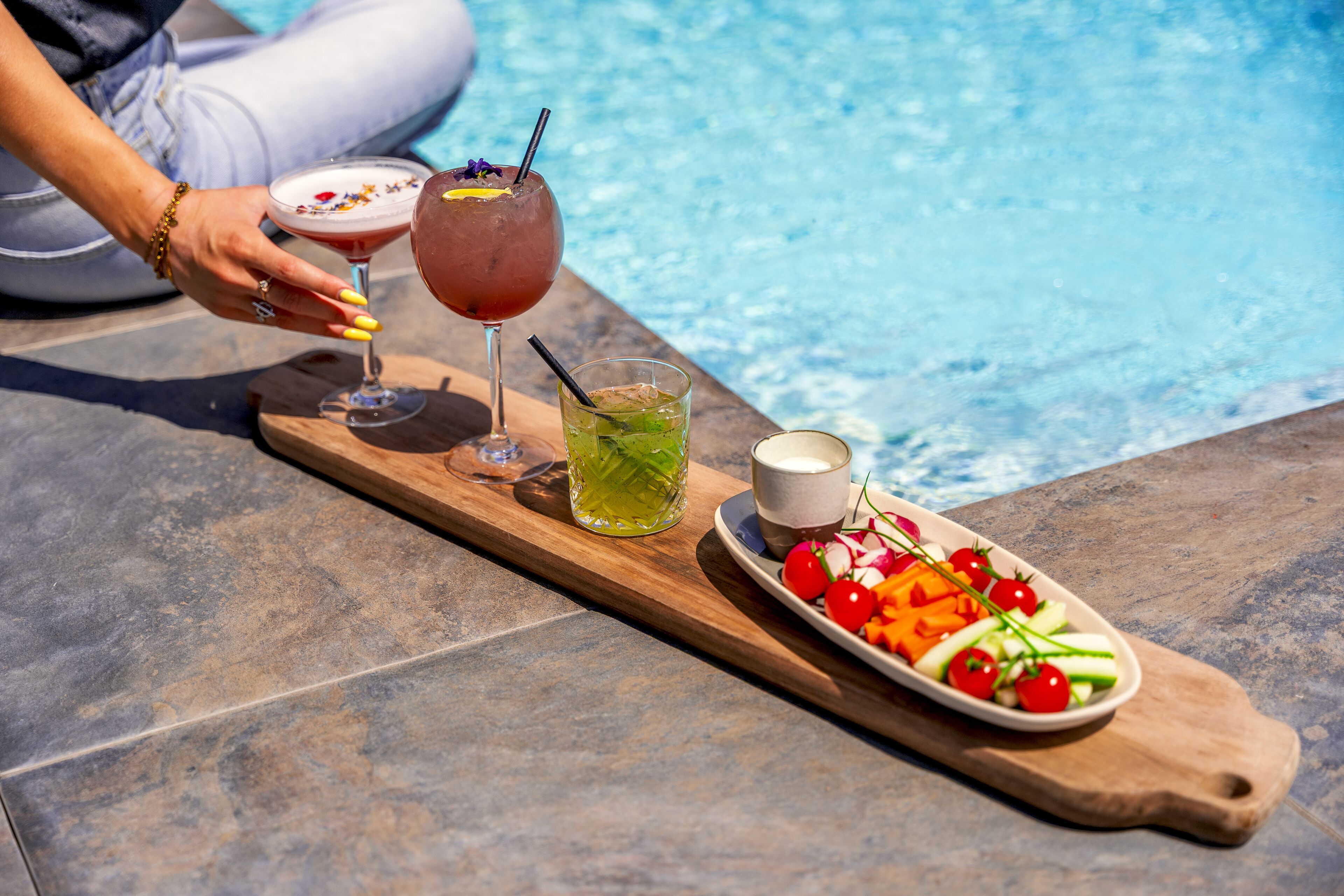 A wooden tray with drinks and a plate of fresh vegetables by a pool.