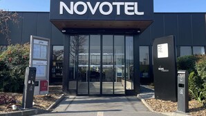 Entrada moderna de un hotel Novotel con fachada de vidrio transparente, un alero de madera y un estacionamiento visible.