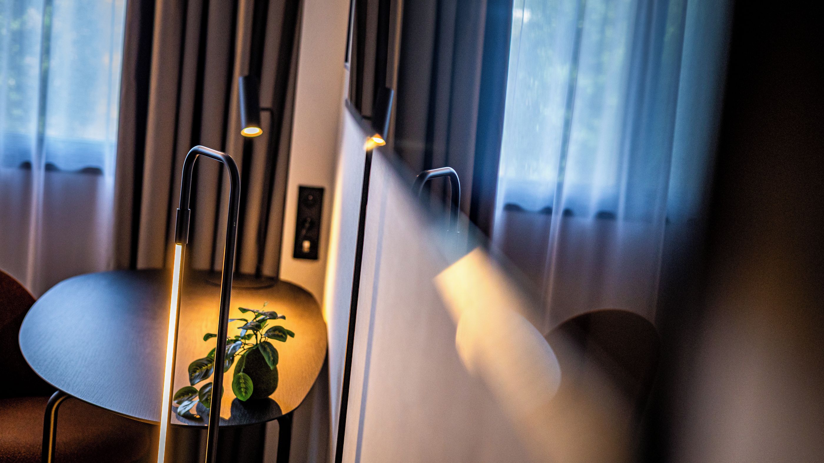 A room with a chair, a table, and a plant. There is a window with curtains and a light fixture.