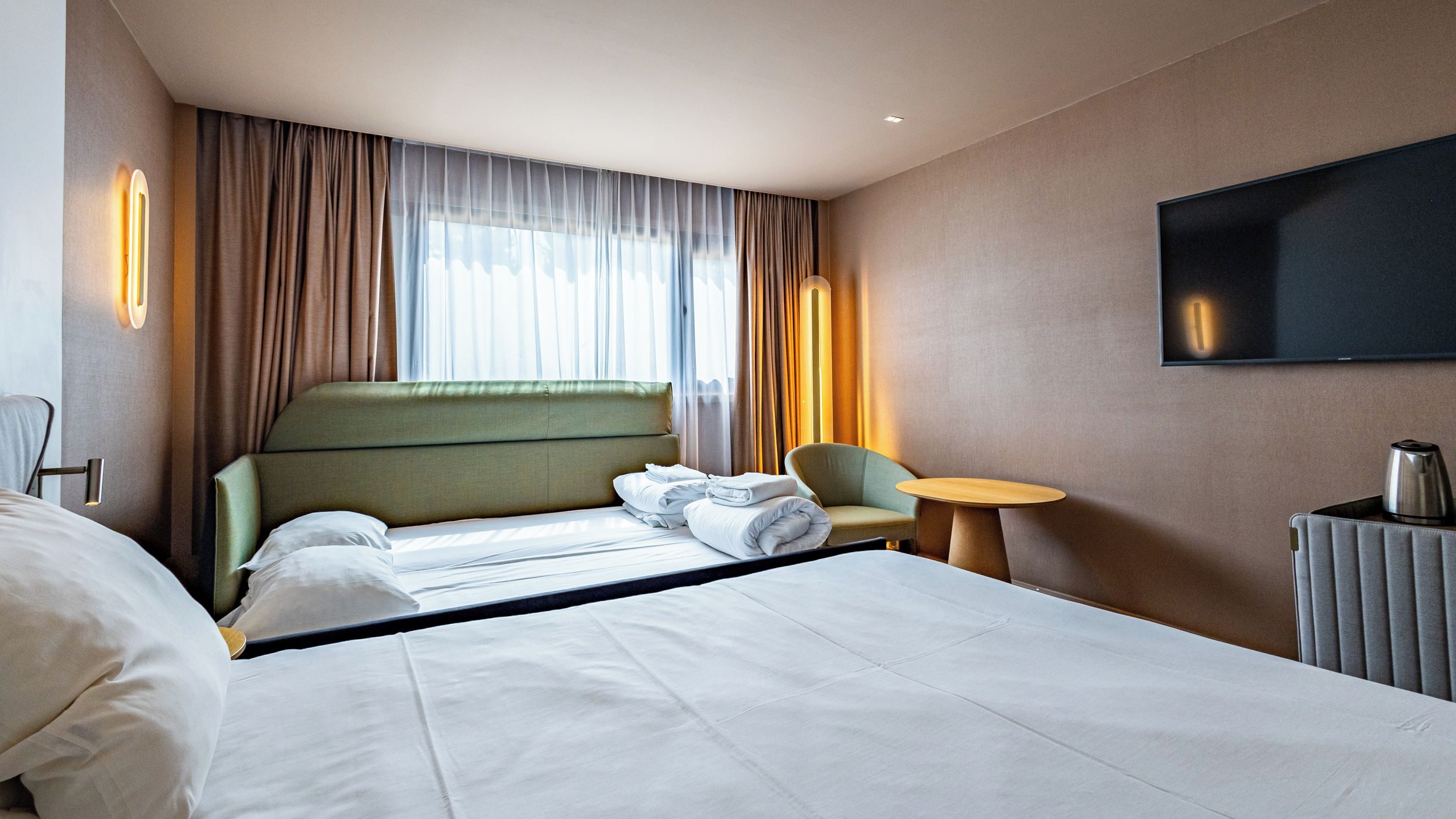 A hotel room with a large bed, a chair, a small table, and a TV mounted on the wall.