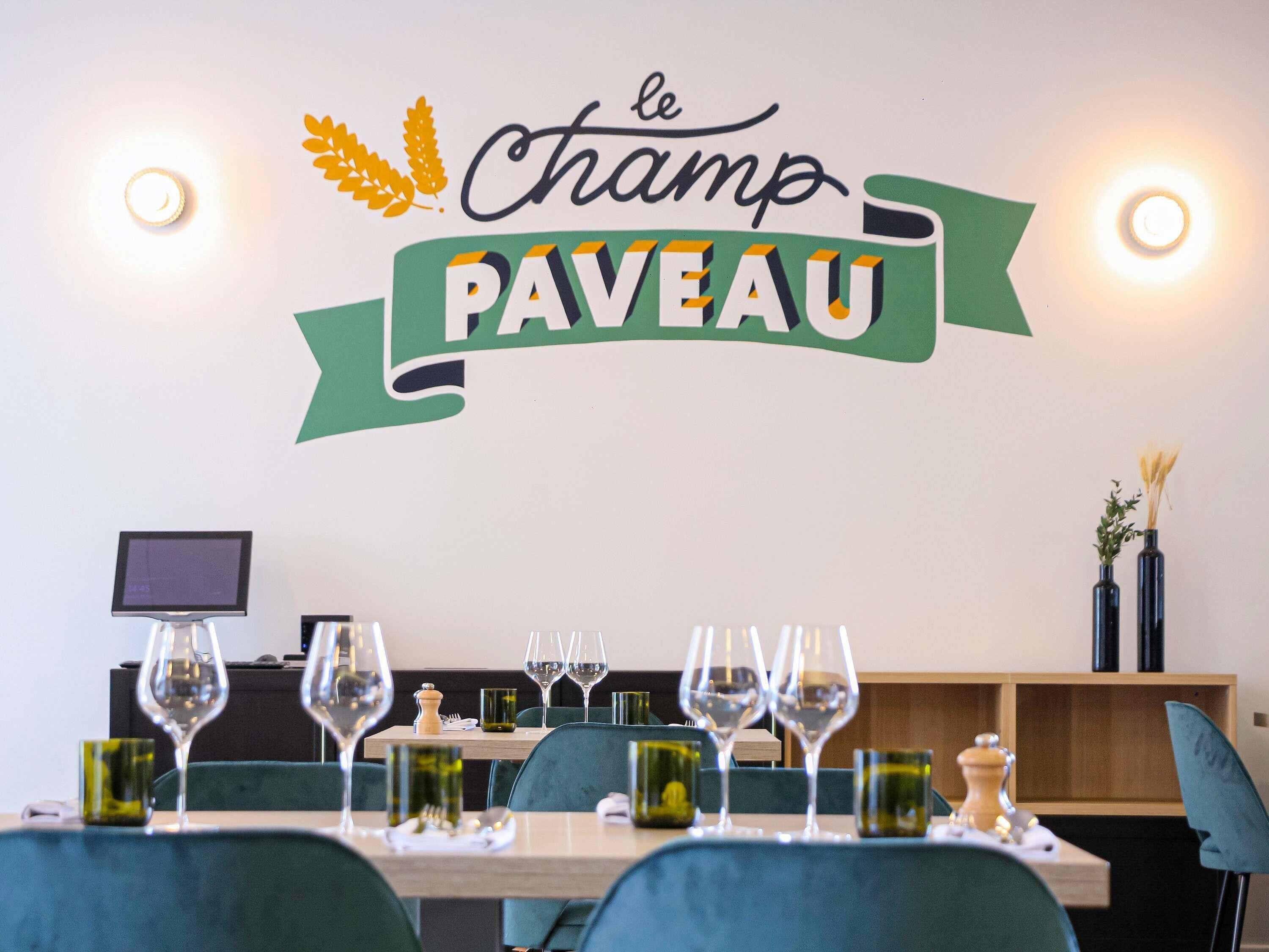 A restaurant interior with a wall sign reading 'le Champ PAVEAU,' a dining table set with wine glasses, and a small screen on the wall.