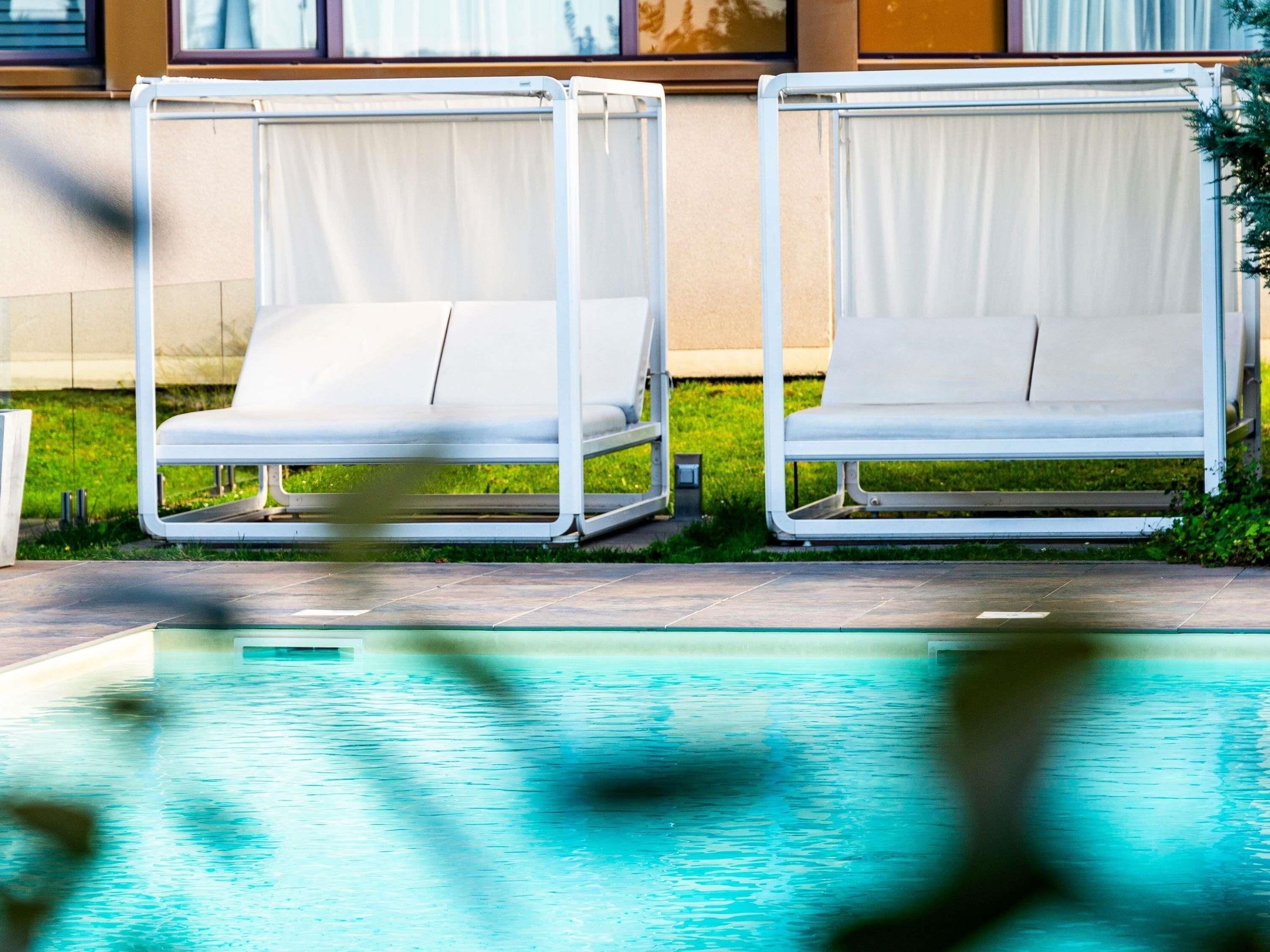 Two white lounge chairs with metal frames, positioned on a tiled patio next to a swimming pool.