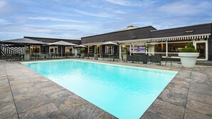 Una piscina con solárium azulejado, mobiliario exterior y un edificio con zona de bar al fondo.