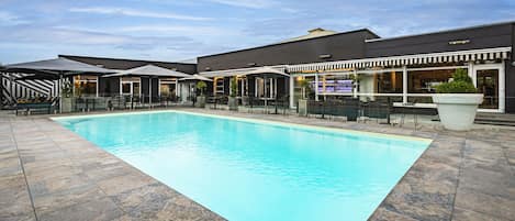 Ein Schwimmbecken mit einer gefliesten Liegefläche, Sitzgelegenheiten im Freien und ein Gebäude mit Barbereich im Hintergrund.