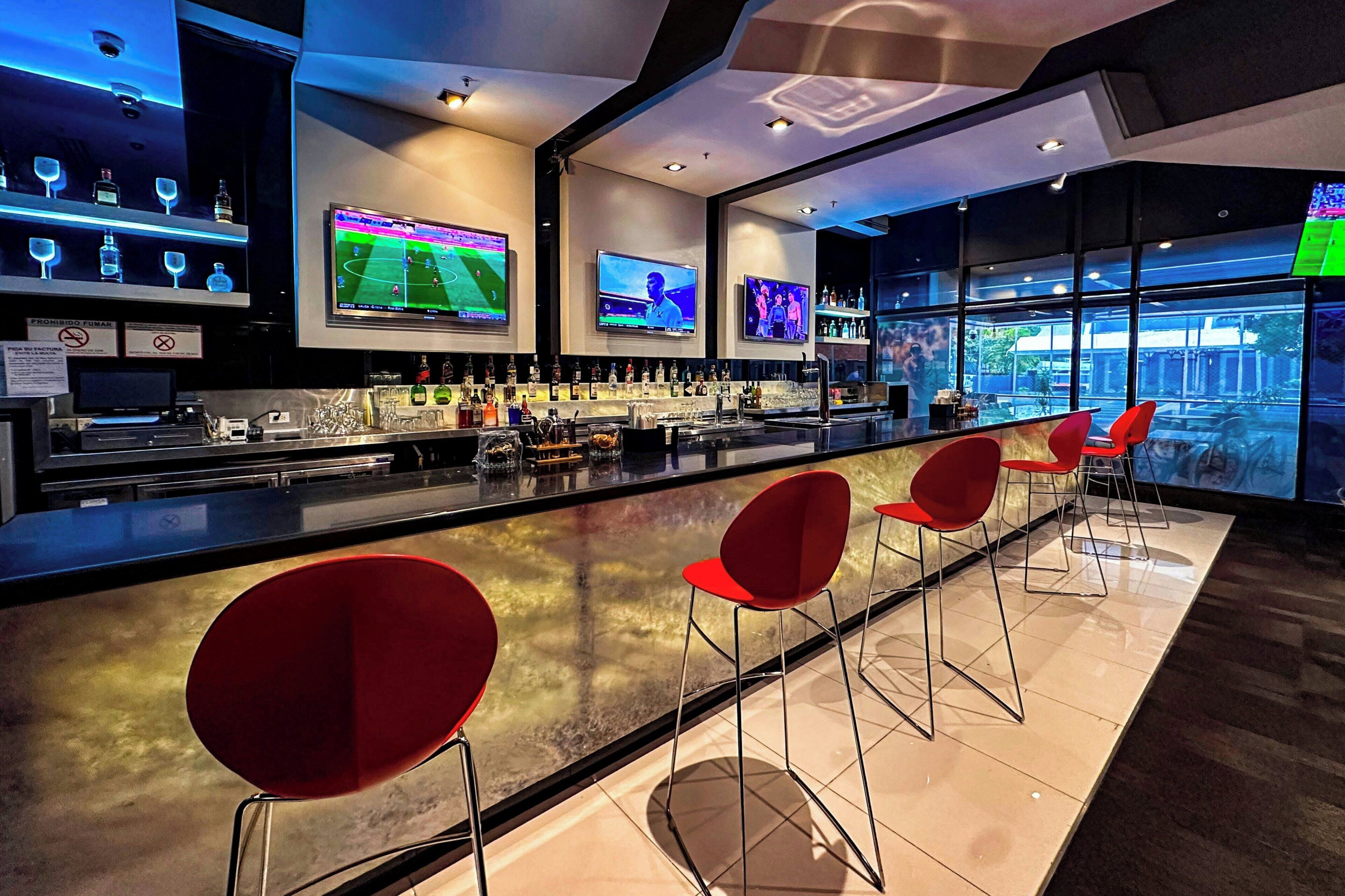 Et moderne barområde med flere fjernsyn, der viser sportsbegivenheder, en velassorteret bar og røde barstole.