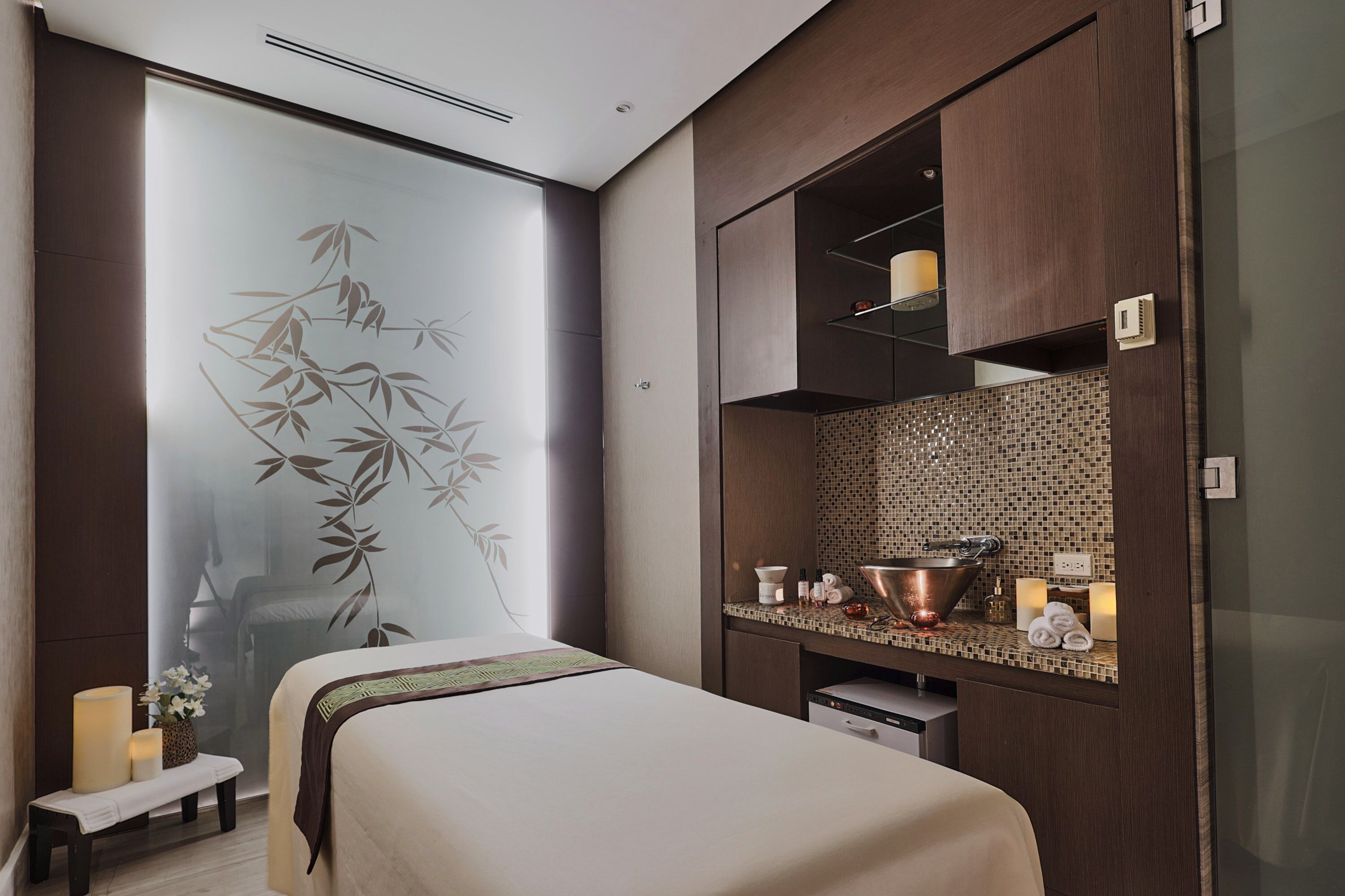 A modern hotel room with a bed, a small bench, a wall with a bamboo design, a bathroom with a sink, and a mirror.