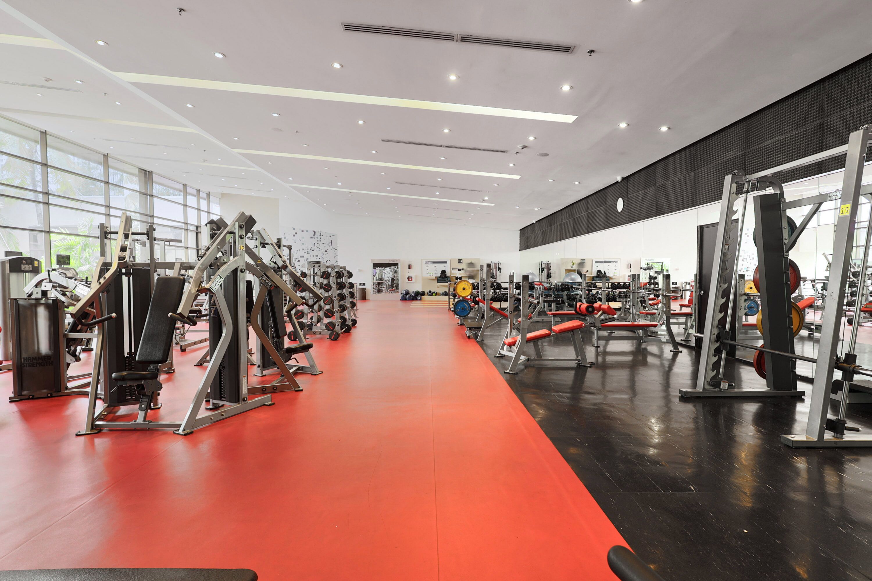 Et fitnesscenter med forskellig træningsudstyr, rødt gulv og store vinduer.