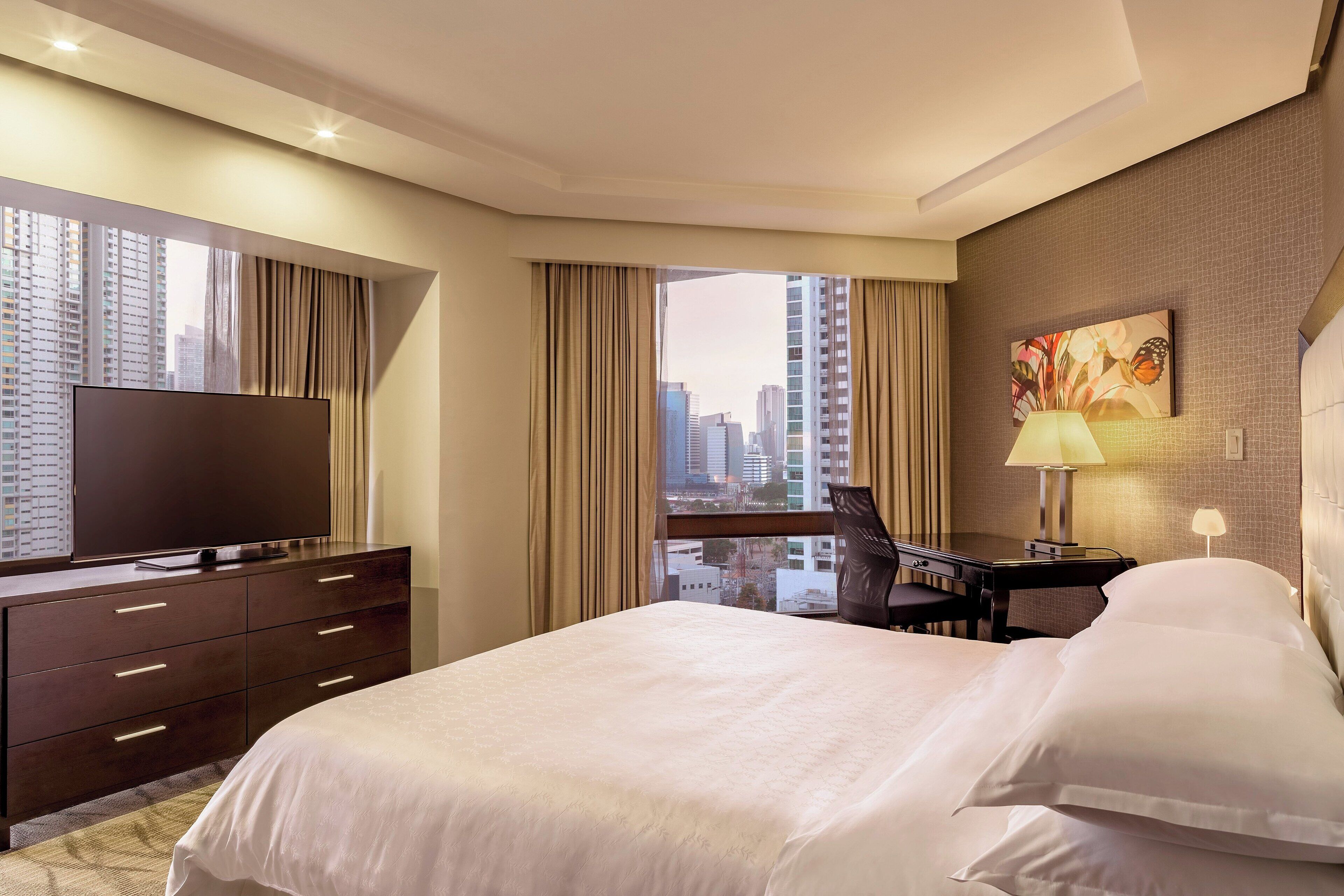 Habitación de hotel con una cama grande, un escritorio, una silla, un televisor y vistas a la ciudad.