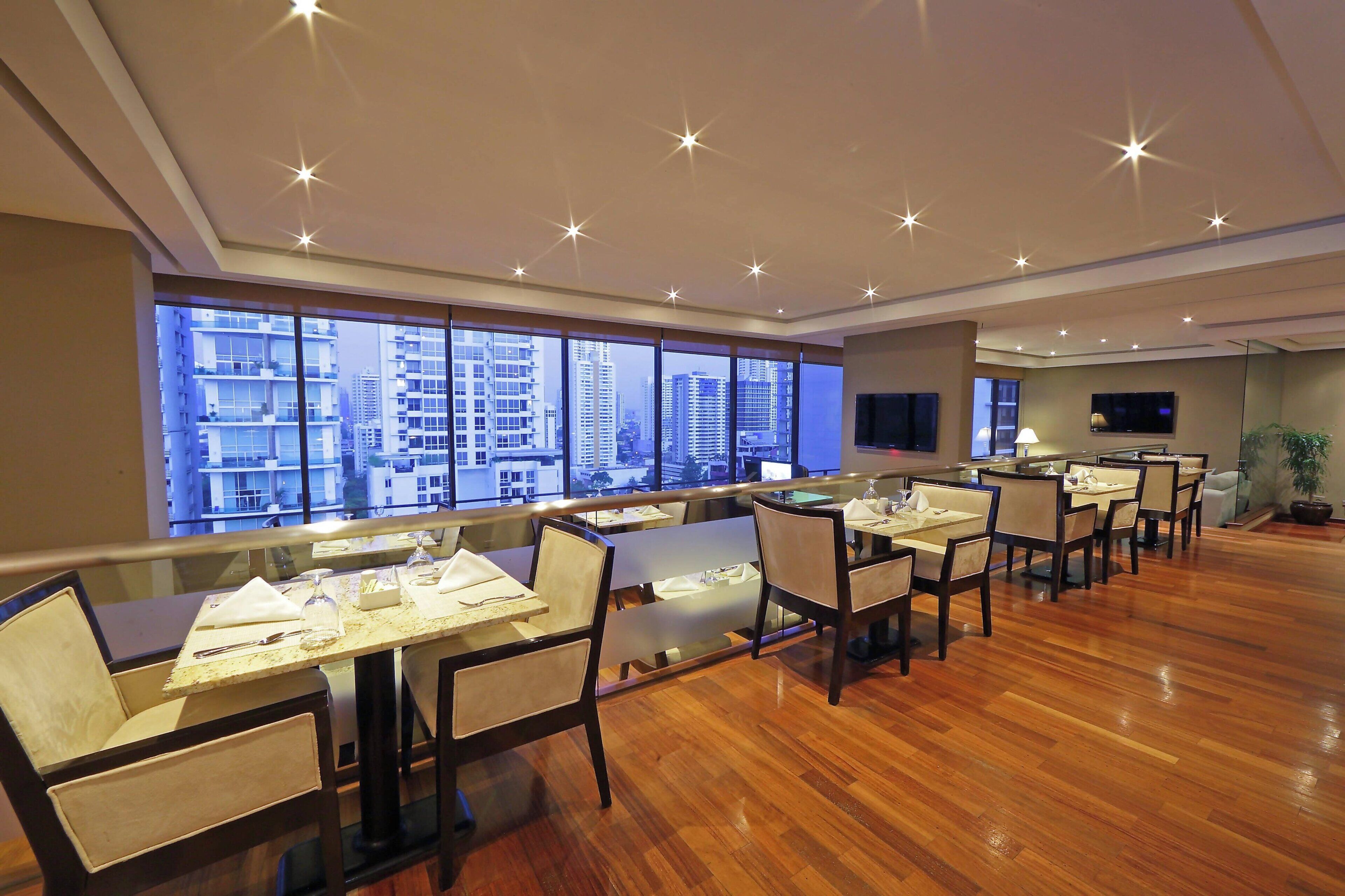 A modern dining area with multiple tables and chairs, large windows with a city view, and a flat-screen TV mounted on the wall.