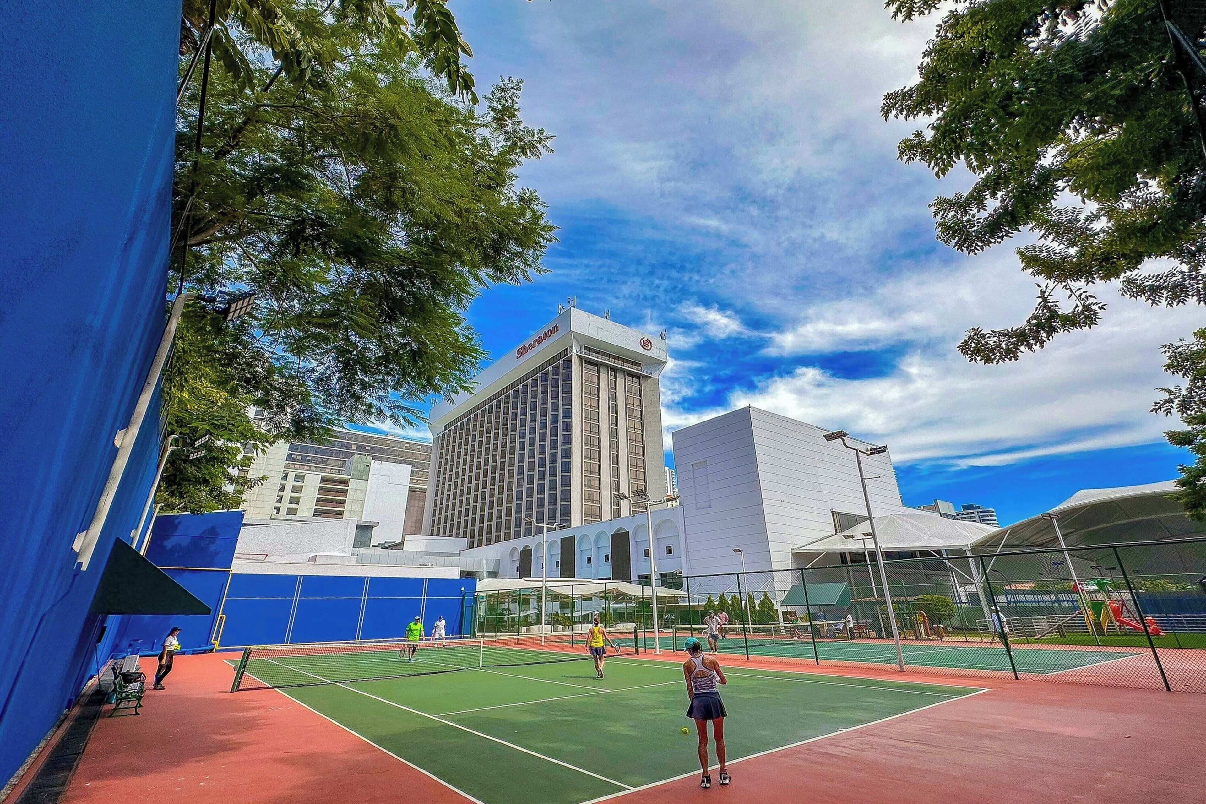 An outdoor tennis court with a net, surrounded by trees and buildings.