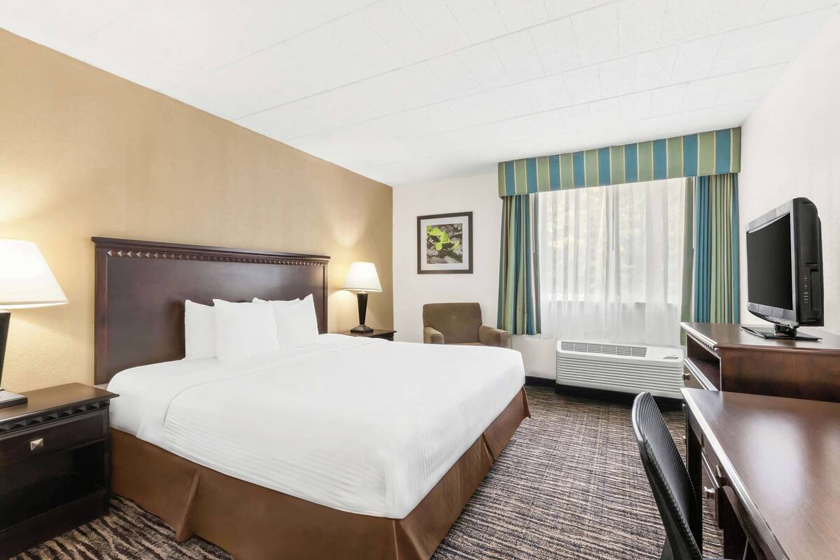 A hotel room with a large bed, two bedside tables with lamps, a chair, a desk with a television, and a window with curtains.