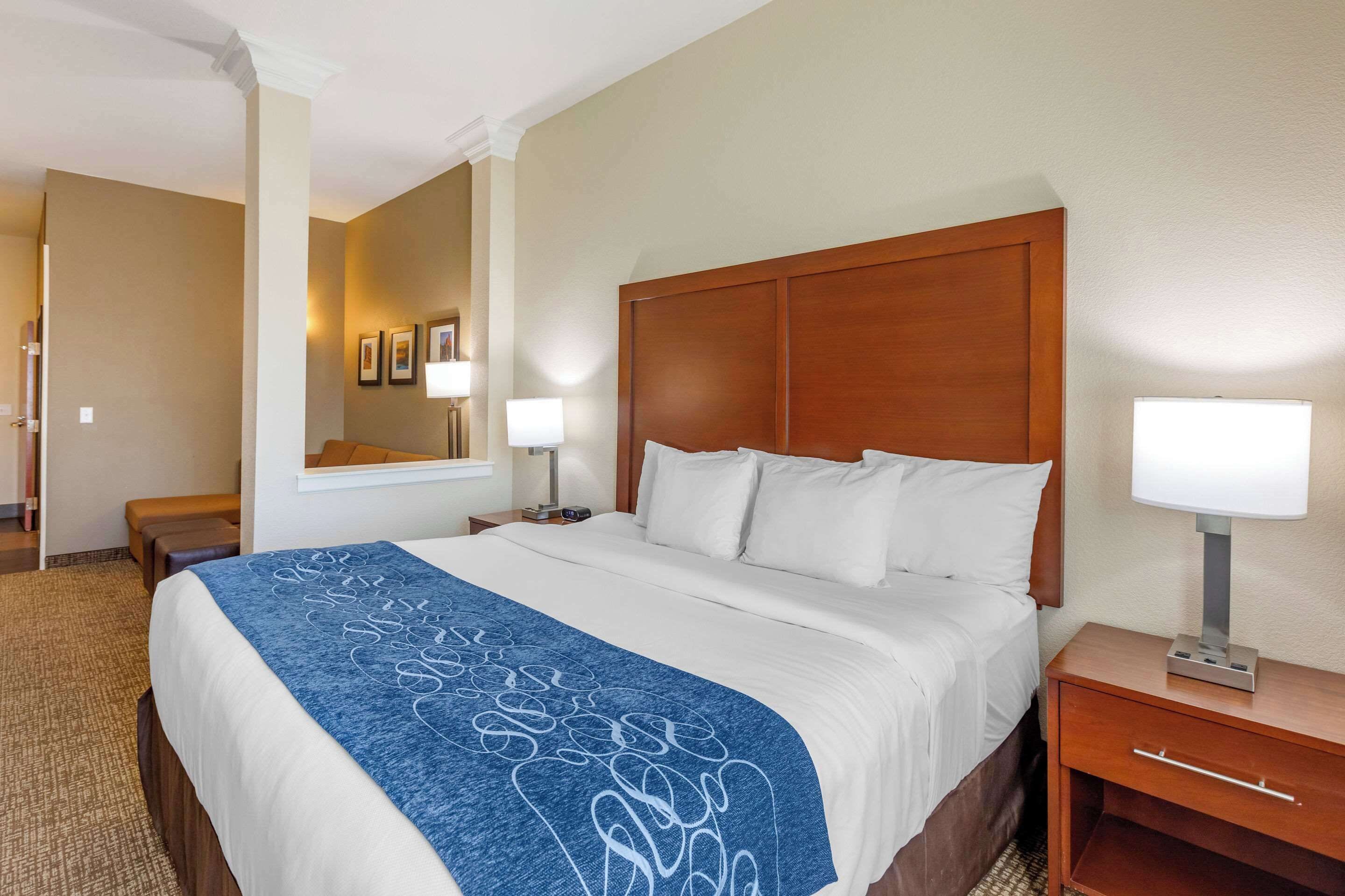 A hotel room with a large bed, a wooden headboard, a nightstand with a lamp, a bench, and a mirror on the wall.
