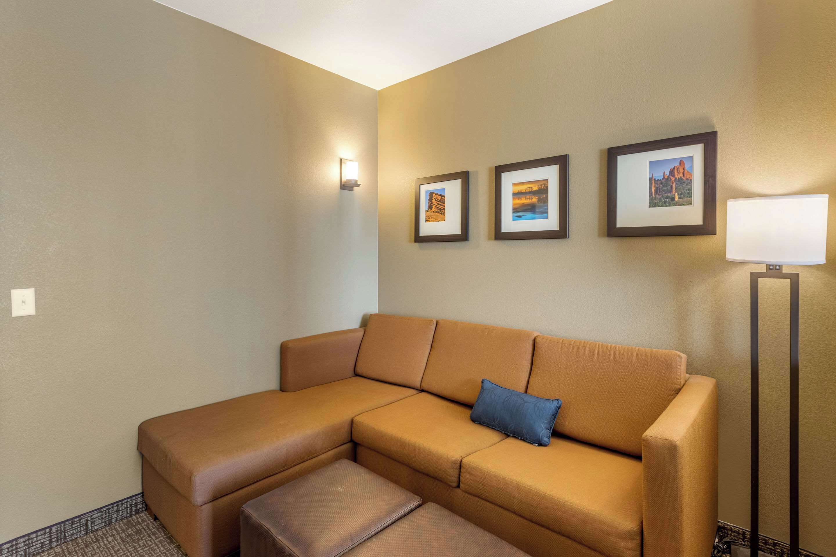 A corner living room with a tan sectional sofa, a small ottoman, and three framed pictures on the wall.