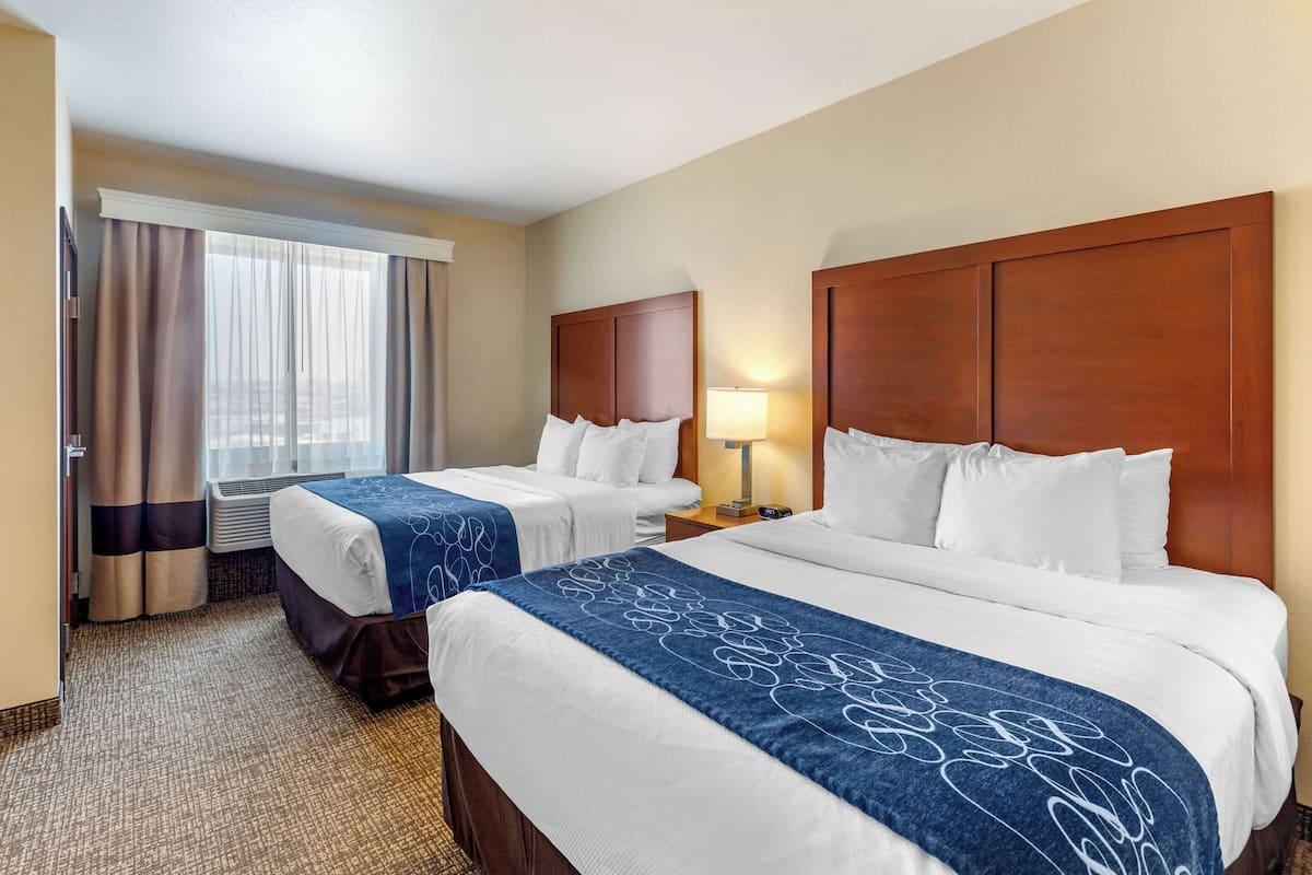 A hotel room with two beds, a large window with curtains, a nightstand with a lamp, and an air conditioning unit.