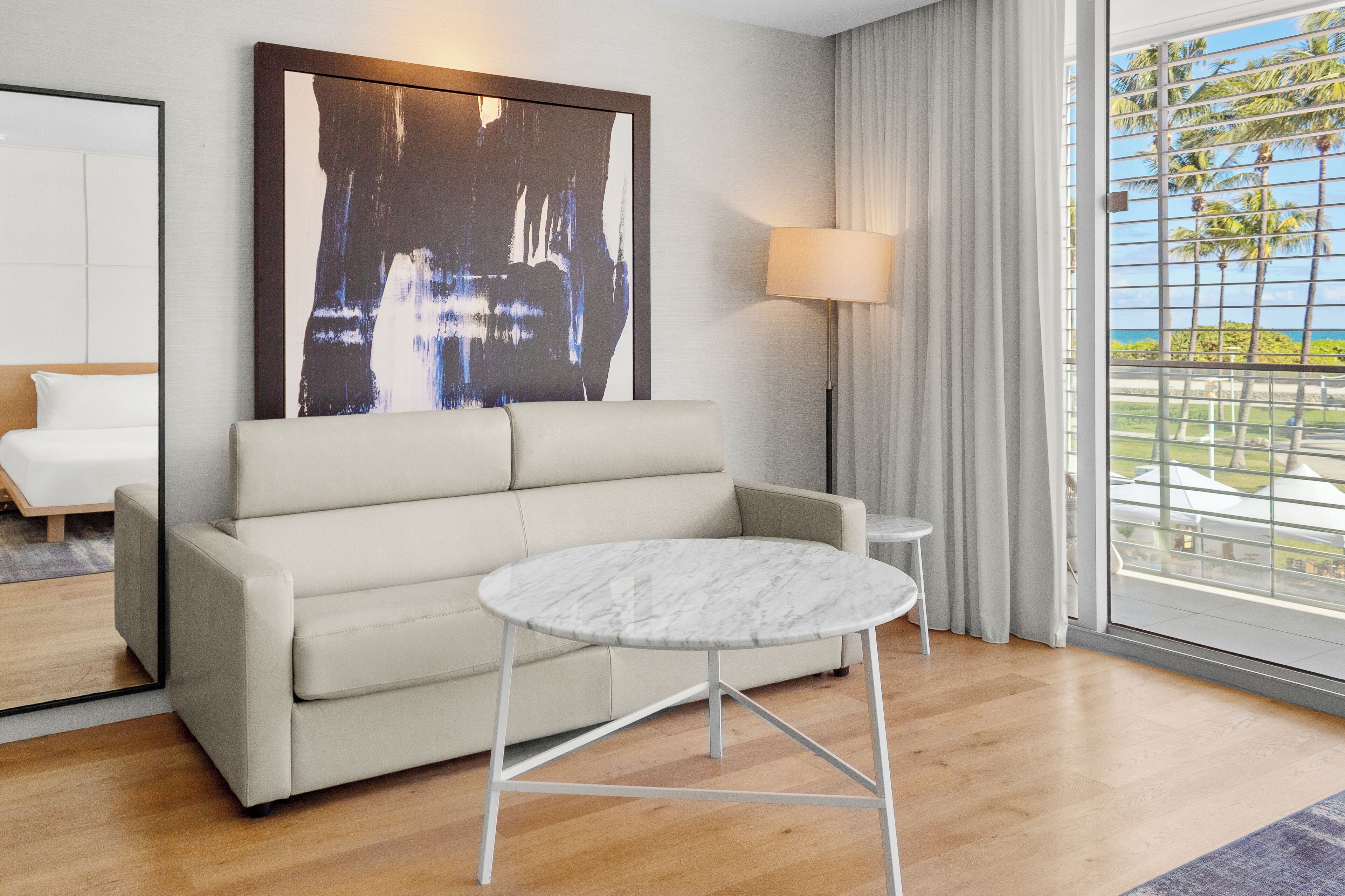 A modern hotel room with a beige sofa, a marble coffee table, a large abstract painting, a floor lamp, and a view of palm trees and the ocean through the window.