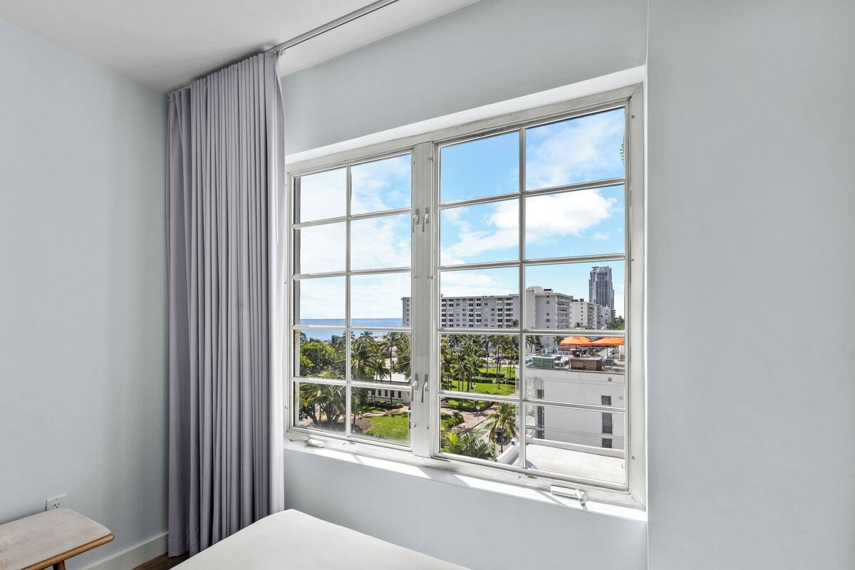 Ein Zimmer mit einem großen Fenster, einer Bank und Blick auf eine Stadtlandschaft sowie Palmen.
