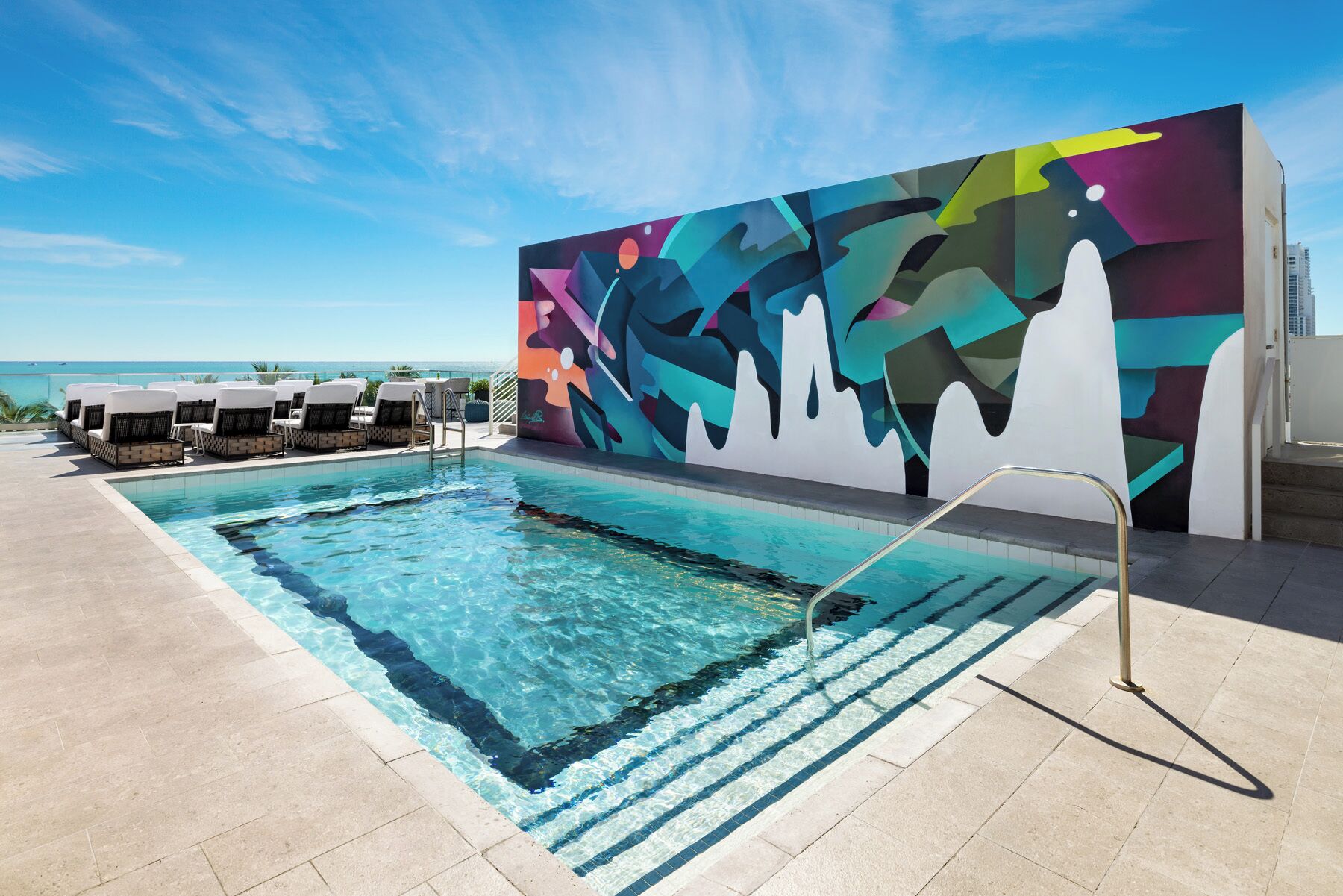 Un espace piscine agrémenté d’une fresque murale colorée, de chaises longues et offrant une vue dégagée sur l’océan.