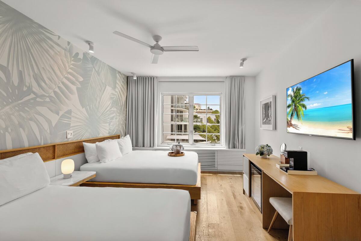 Ein Hotelzimmer mit zwei Betten, einem Schreibtisch, einem Deckenventilator, einem Fenster mit Blick auf ein Gebäude und einem Bild von einem Strand.