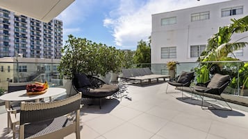 A rooftop terrace with a glass balustrade, outdoor seating, and a view of a modern building.